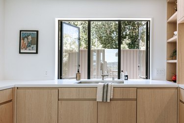 Light wood kitchen cabinets with white countertop and built-in shelving. Tri black framed windows and framed portrait.